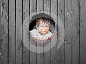 Child looks out of a round wooden window and smiles. The boy climbed out of the round window and smiles pretty straight into the