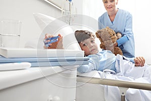 Child lying in bed in hospital room taking a pair of glasses with a nurse holding a teddy bear behind him