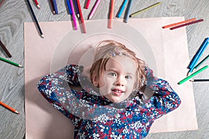 Child lying on the floor paper looking at the camera near crayons. Little girl painting, drawing. Top view. Creativity concept.