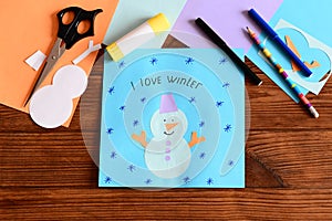 Child made a funny paper snowman applique, drew a snowflake and wrote I love winter. Stationery, colored paper sheets on a table
