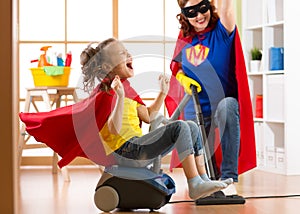 Child and mother dressed as superheroes using vacuum cleaner in room. Family middle-aged woman and daughter have a fun