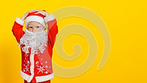 Child in New Year`s Santa Claus costume and white fake beard grabbed his head, copy space, yellow isolated background.