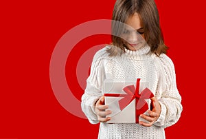 Child opening gift box with red ribbon and looking inside with curiosity on red background