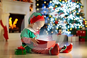 Child opening present at Christmas tree at home. Kid in elf costume with Xmas gifts and toys. Little baby boy with gift box and