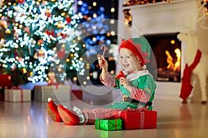 Child opening present at Christmas tree at home. Kid in elf costume with Xmas gifts and toys. Little baby boy with gift box and