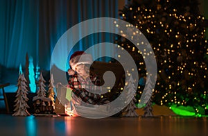 Child opening presents on Xmas eve. Kid in santa hat on front of night Christmas tree home background on Christmas Eve