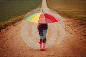 Child in orange rubber boots holding colorful umbrella under rain in autumn. Back view