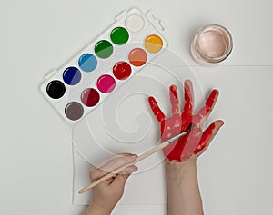 Child painting her hand with red paint and paintbrush. Finger painting or art therapy for children