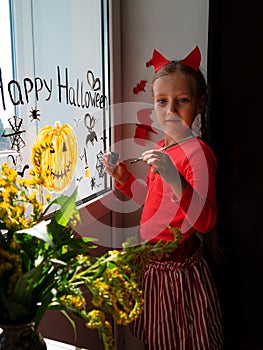 Child painting pumpkin on window preparing celebrate Halloween. Little kid draws decorates room interior with paper bats photo