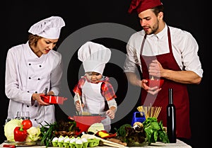 Child with parents cooking at kitchen. Happy family in chef uniform preparing dinner together.