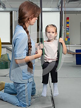 Child with physiotherapist on swing during sensory integration session. Little girl in rehabilitation centre doing