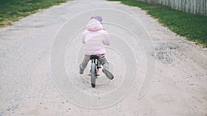 Child in pink jacket and lilac hat rides balance bike on dirt road, in countryside