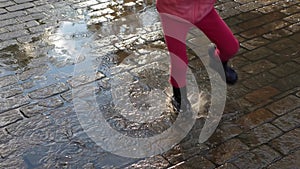 Child in pink jumps on puddles, splashes scatter from rubber boots, slow motion.