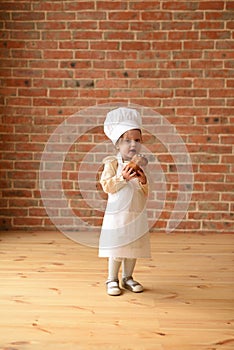 Child playing chef. Cute toddler girl in chef apron and hat eating sweet bun at brick wall in empty room