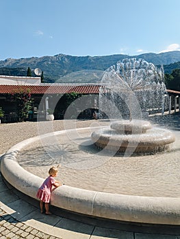 The child is playing next to the fountain. A little girl in a pink dress wets her hands near a large fountain in Budva