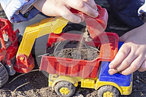 The child is playing in the street with sand; he loads the earth in an dump truck toy