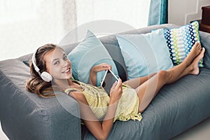 Child playing tablet and listening to music in head phones while relaxing on couch in living room at home