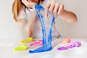A child plays with colored slime at a table in the room. The girl holds in her hand and stretches the blue slime to transparency