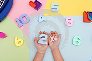 The child plays with multi-colored numbers. Child`s hands hold numbers top view on colored pastel background