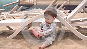 The child plays sits on the sand. A little boy on the beach near the river plays with his feet in the sand near the sun