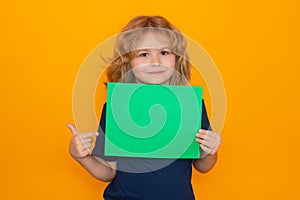 Child pointing on empty green sheet of paper, isolated on yellow background. Portrait of a kid holding a blank placard