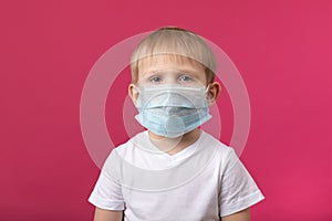 Child preschooler boy closeup shot in medical mask looking at the camera on a pink background