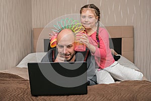 A child prevents a man from working on a laptop in his bedroom. The daughter puts the slinky toy to the father`s head. Family is