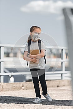 child in protective mask standing with bag and book on bridge air