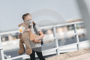 child in protective mask walking with teddy bear and book on bridge air