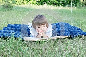 Child reading book lying on stomach outdoor, smiling cute little girl, children education and development