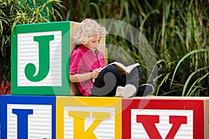 Child reading book in school yard. Kid learning abc letters. Little boy sitting on wooden toy blocks with alphabet in preschool or