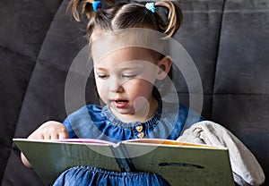 Child reading. Little cute girl 1-3 is reading a book with a toy plush hare sitting on a sofa