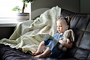 Child reading. Little cute girl 1-3 is reading a book with a toy plush hare sitting on a sofa