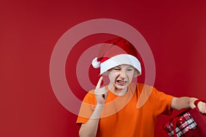 A child on a red background in a Santa Claus hat is holding a gift bag in his hands, grimacing and looking at him