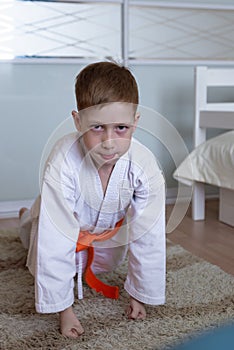 Child red-haired boy is engaged in karate, training push-ups at home in the form of a kimono with an orange belt