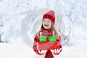 Child in red hat with Christmas presents and gifts in snow. Winter outdoor fun. Kids play in snowy park on Xmas eve. Little girl