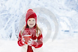 Child in red hat drinking hot chocolate in snow on Christmas vacation. Winter outdoor fun. Kids play in snowy park on Xmas eve.