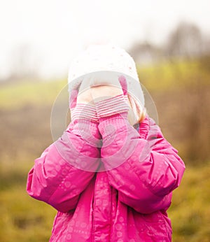 A child in a red jacket and a white hat plays hide and seek on the street. Autumn. The face is covered with mittens.