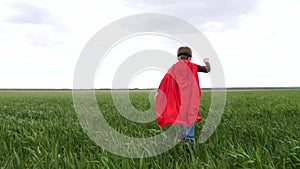 A child in a red raincoat runs on green grass against a blue sky. The boy is playing a superhero.