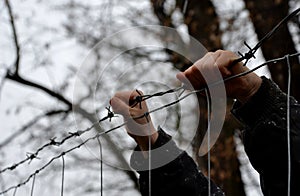 Child in a refugee camp behind a wire fence in winter rainy day. holding barbed wire with small hands. knitted gloves white finger