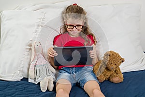 Child is resting at home. Little girl 6, 7 years old sitting in bed with toys, looking at her tablet, smiling