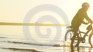 Child rides a bicycle on the water in the rays of the setting sun along the beach. Sports activities at sunset.