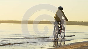 Child rides a bicycle on the water in the rays of the setting sun along the beach. Sports activities at sunset.