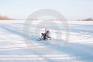 Child rides snowcat on snowy road. Little girl in pink warm jacket enjoys walk in nature and sledding on frozen river on