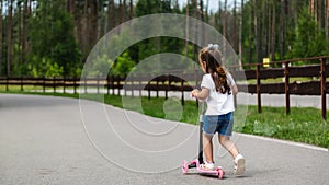 Child riding scooter. Happy little kid girl playing pink kick board on road in park outdoors on summer day, Active children games