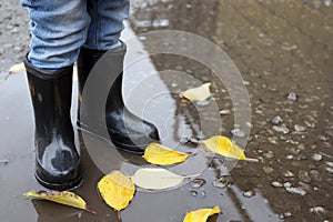 child in rubber boots in autumn puddle after rain with yellow leaves