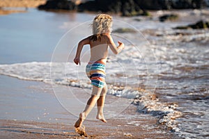 Child running through water close to shore along the sea beach. A boy runs along the sea coast. Rest of children on