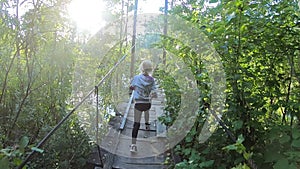 The child runs along the path and reaches the hanging wooden bridge over the river