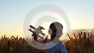 A child runs along the wheat field during sunset, holding a toy plane in his hands. The boy shows the flight of the