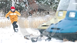 The child runs through the snowdrifts in the direction of a snowmobile in the forest